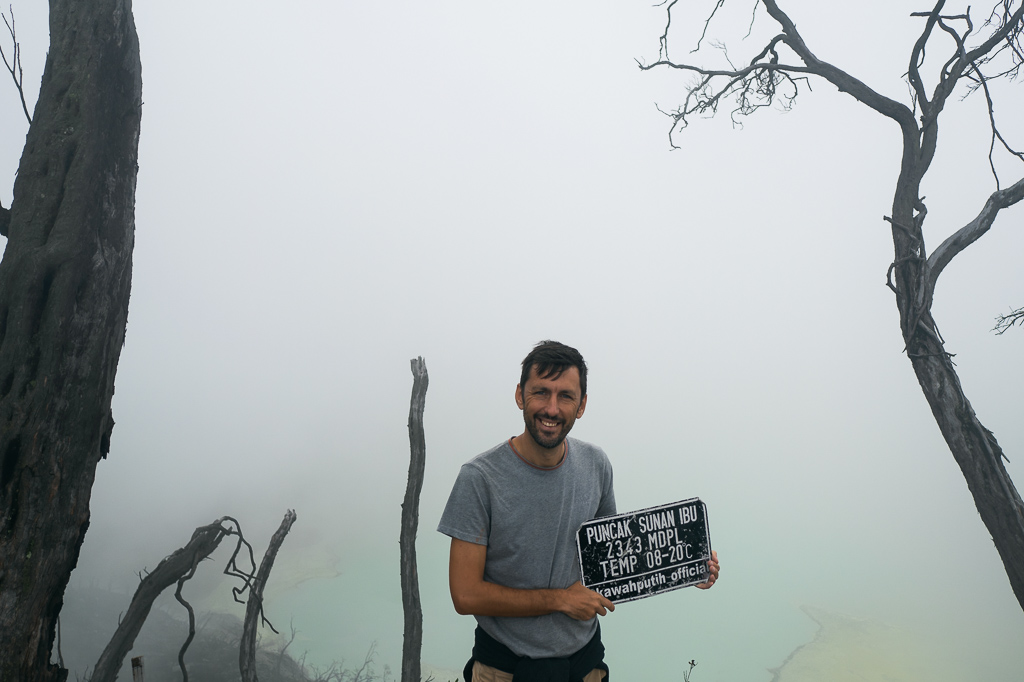Smiling man in gray t-shirt holds a "PUNCAK SUNAN IBU" sign on a foggy, barren Mount Ijen