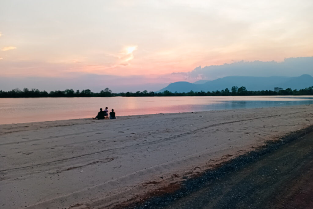 Kampot river beach at sunset with 3 people, calm water, and distant mountains.
