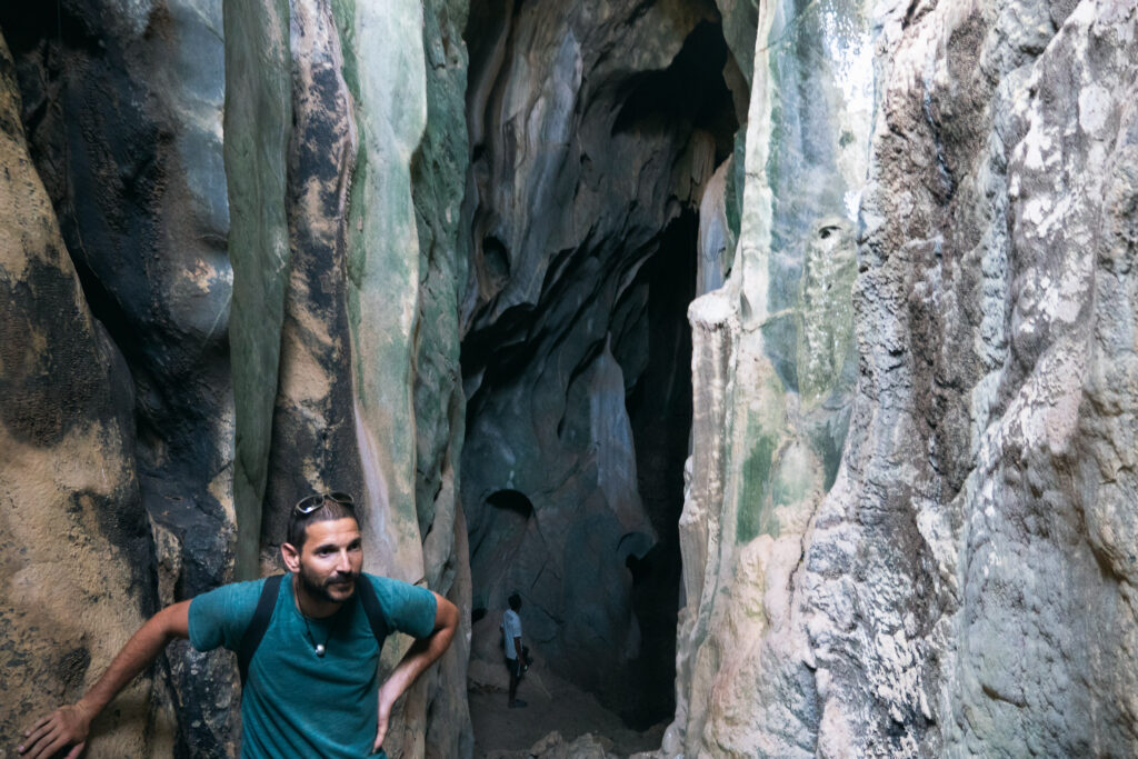 A man standing at a large cave entrance with rugged rock formations in Kampot