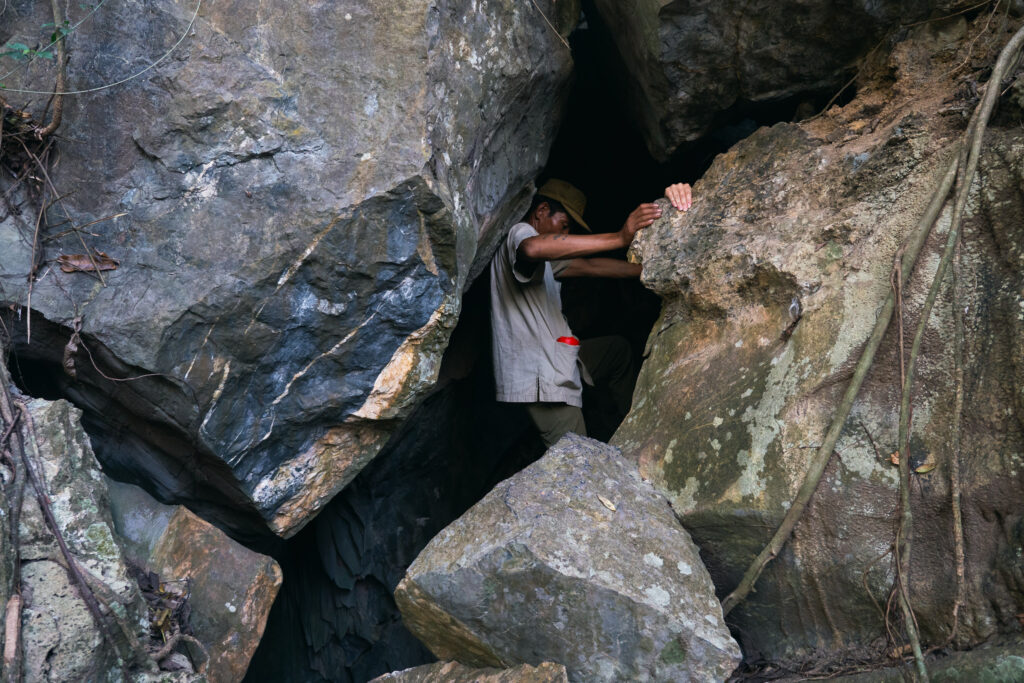 A person navigates a narrow rocky cave entrance, gripping rough rock surfaces
