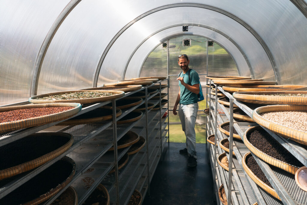 A man stands in a clear arched drying facility, surrounded by shelves of baskets filled with drying beans or grains