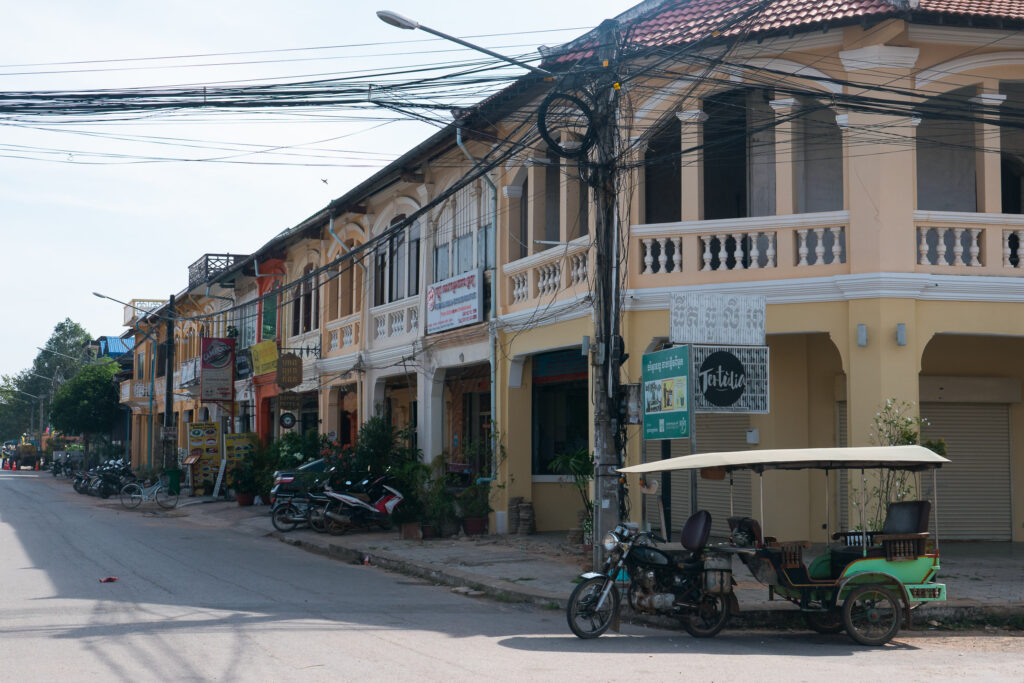 A quiet street in Kampot, Cambodia, lined with pastel French colonial buildings and a few parked motorbikes and a green tuk-tuk