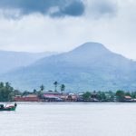 A fishing boat on the calm Preaek Tuek Chhu River in Kampot, Cambodia, with palm-lined banks and a hazy mountain in the background