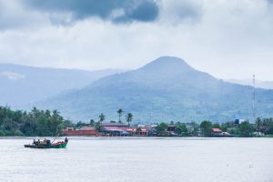 A fishing boat on the calm Preaek Tuek Chhu River in Kampot, Cambodia, with palm-lined banks and a hazy mountain in the background