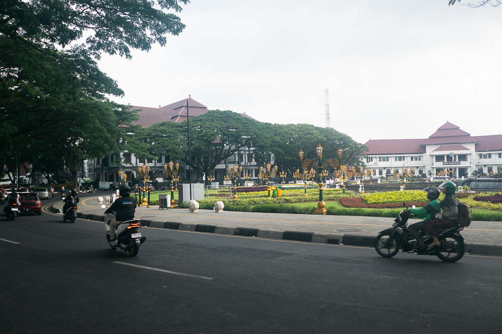 Street scene in Malang, Indonesia, featuring motorcyclists riding past a landscaped city square with colonial-era buildings in the background