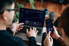 Two pairs of hands look at investing numbers on a computer and phone.