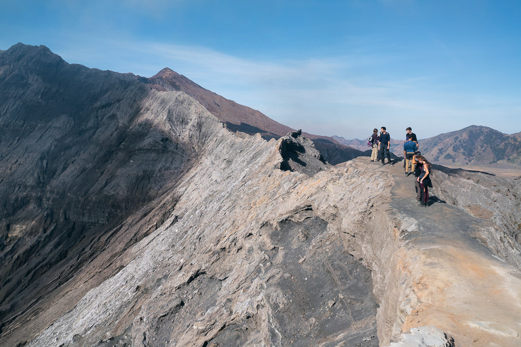 A group of hikers walks along a narrow, rugged volcanic ridge on Mount Bromo