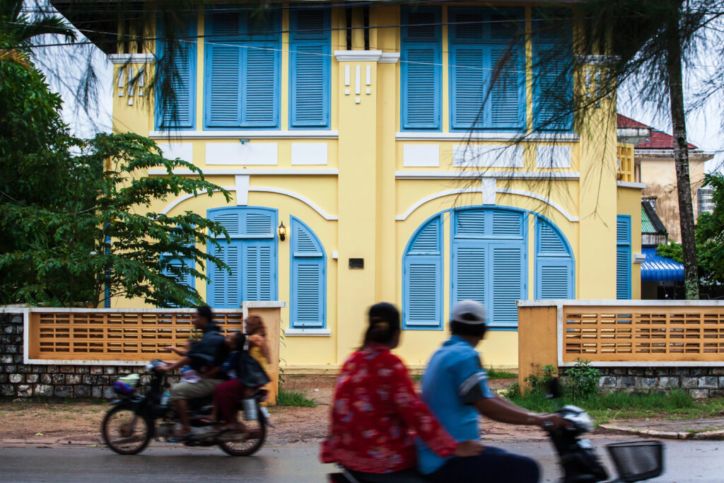Khmer locals ride motorcycles past old French colonial buildings in Kampot, Cambodia.