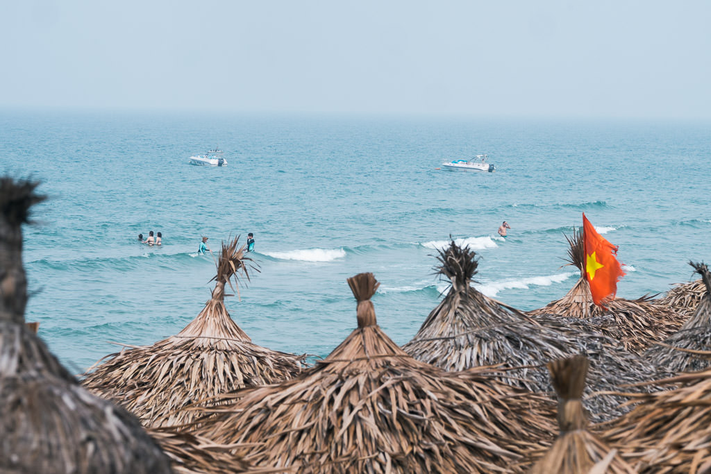 People swimming in and boats on the sea and the roof of the huts by the sea in Hoi An, Vietnam.