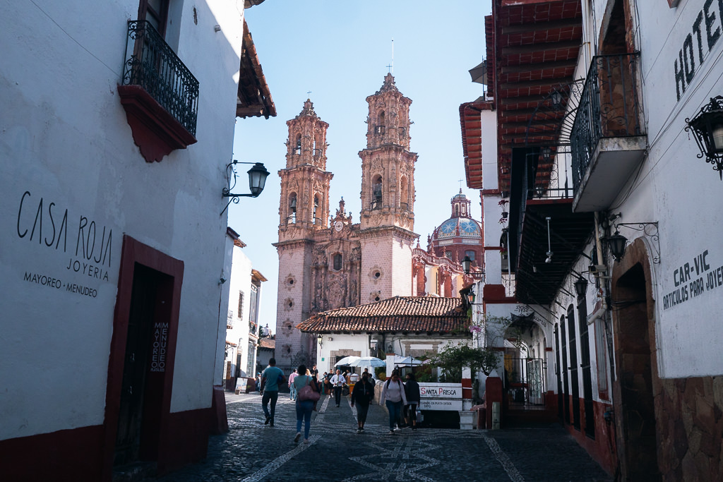A street view in Taxco, Mexico, shows a cobblestone street lined with white buildings and Santa Prisca Church in the background