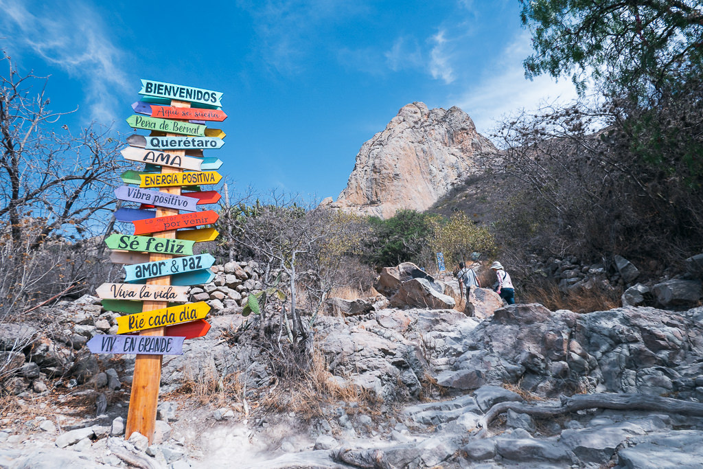 A signpost with several directional arrows stands in front of the towering Peña de Bernal monolith near Querétaro, Mexico.