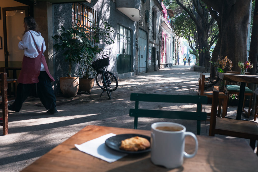 A street view in Roma Norte, Mexico City, with a white mug of coffee and a cookie on a wooden table in the foreground.