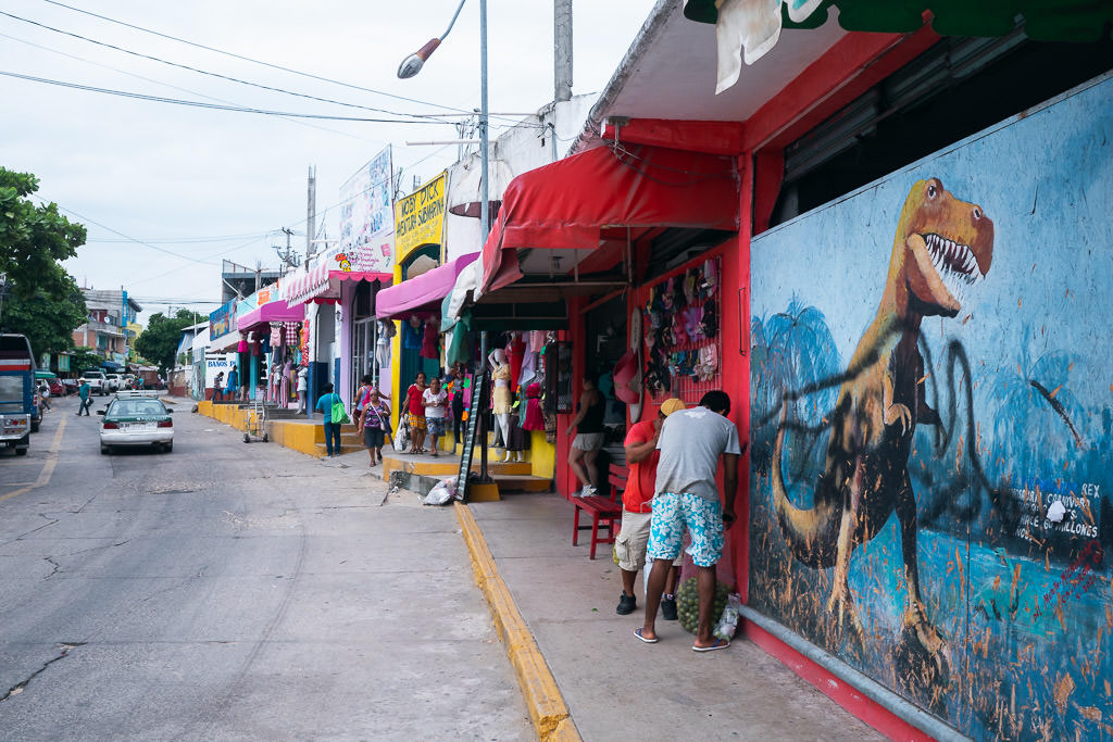 A street scene in Puerto Escondido featuring colorful storefronts, a prominent dinosaur mural on a red building, and people walking along the sidewalk.