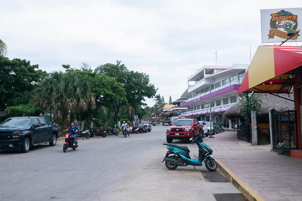 Street scene in Zicatela, Oaxaca, on an overcast day with scooters, pickup trucks, colorful buildings, and lush greenery.