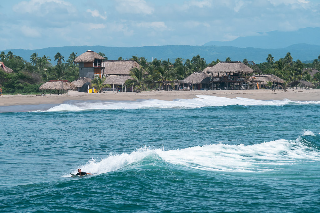 Wide shot of a surfer paddling into a wave at Chacahua Beach in Oaxaca, with rustic beachfront cabanas, palm trees, and lush hills in the background.