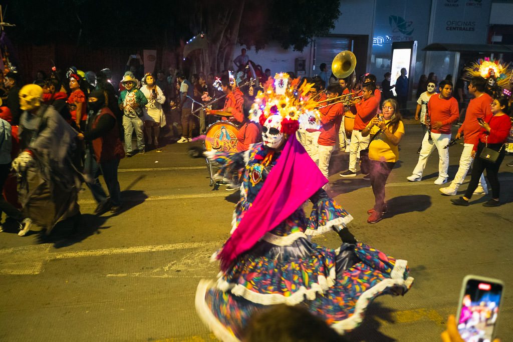 A Day of the Dead parade at night with a dancer in a colorful skirt, pink shawl, and skull mask leads the way, followed by a brass band and participants in festive costumes