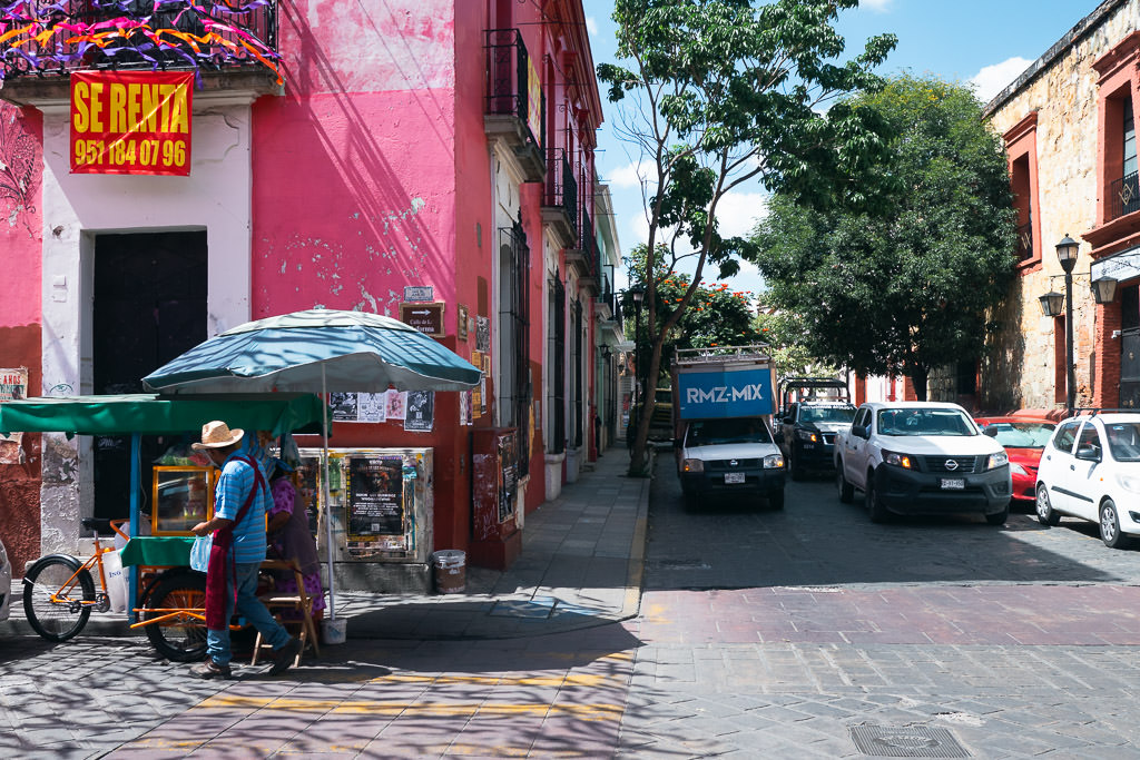 Street view in the Centro area of Oaxaca de Juárez, featuring a pink building, a street vendor cart, and cars passing by.