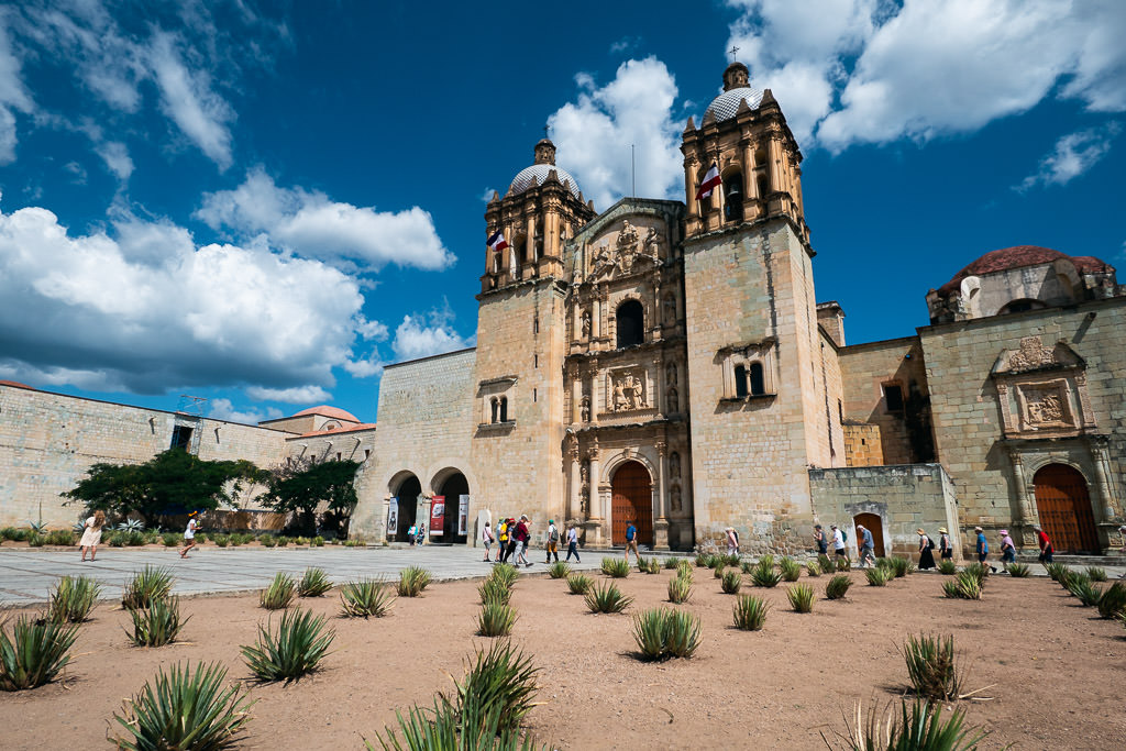 A wide-angle, eye-level shot captures the Baroque facade of the Church of Santo Domingo de Guzmán in Oaxaca, Mexico