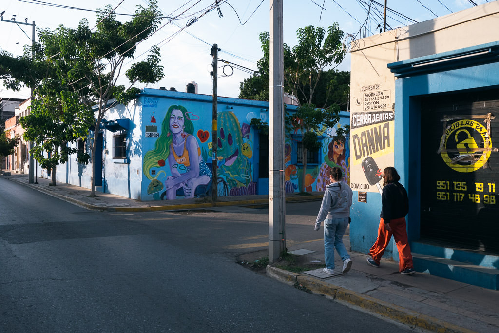 Two pedestrians walk along a sidewalk in Oaxaca, Mexico, past buildings adorned with vibrant murals