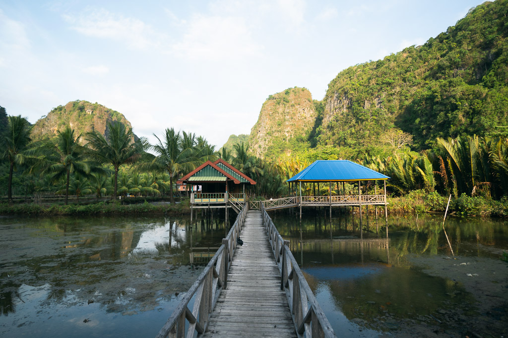 Wooden boardwalk in Rammang-Rammang leading to stilt gazebos over calm water, surrounded by palm trees and limestone karsts