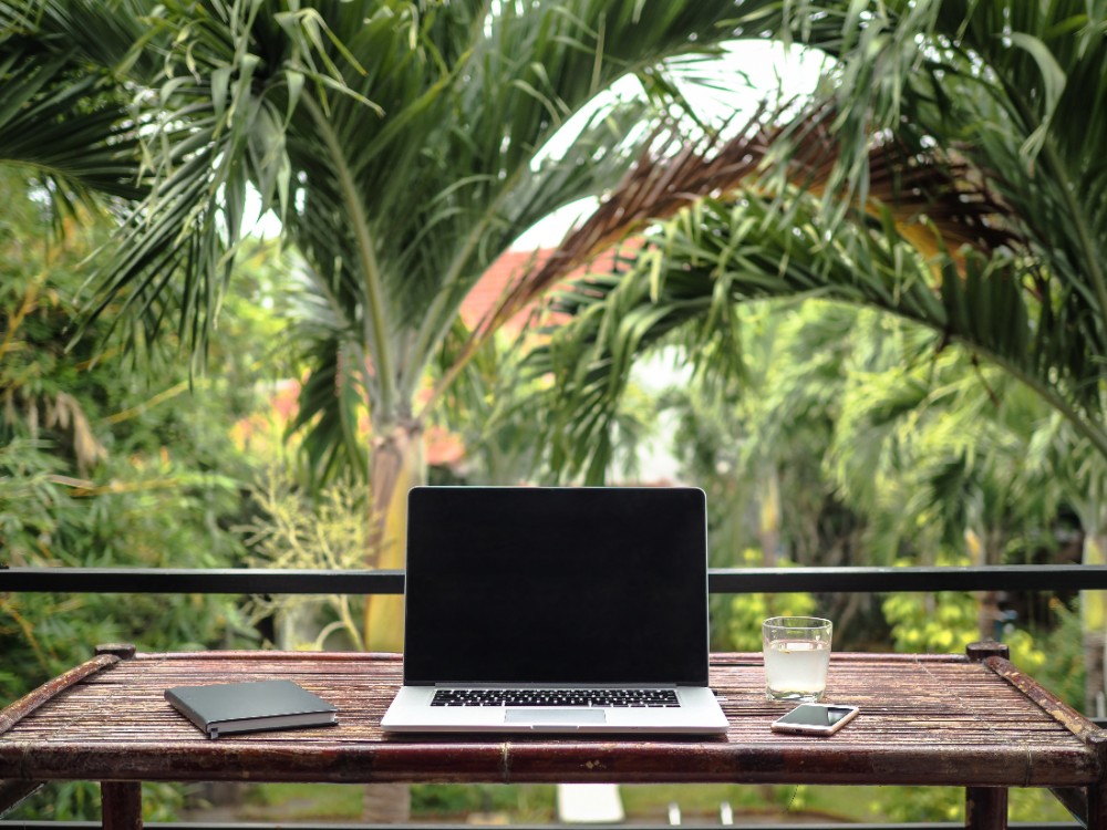 A laptop with a blank screen, a notebook, a smartphone, and a glass of water are arranged on a bamboo table