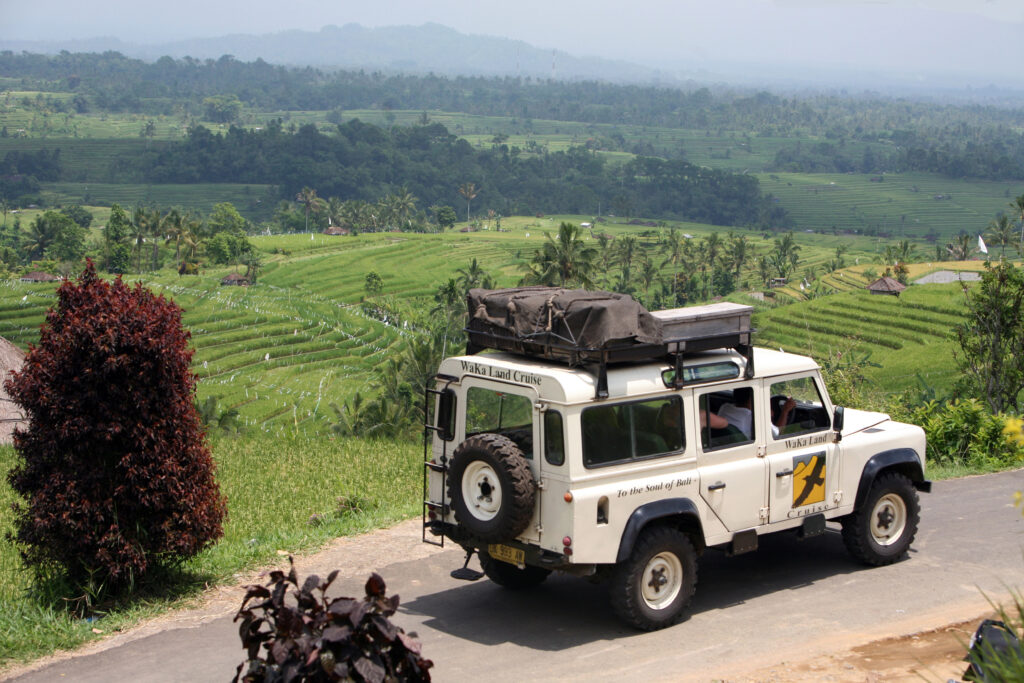 White 4x4 vehicle driving past lush green rice fields in Bali, Indonesia, with hills and palm trees in the background.