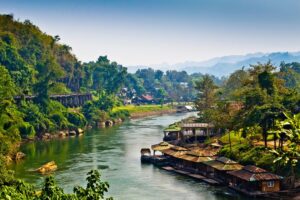 The River Kwai in Thailand surrounded by houses, trees, railroad, and mountains in the background.