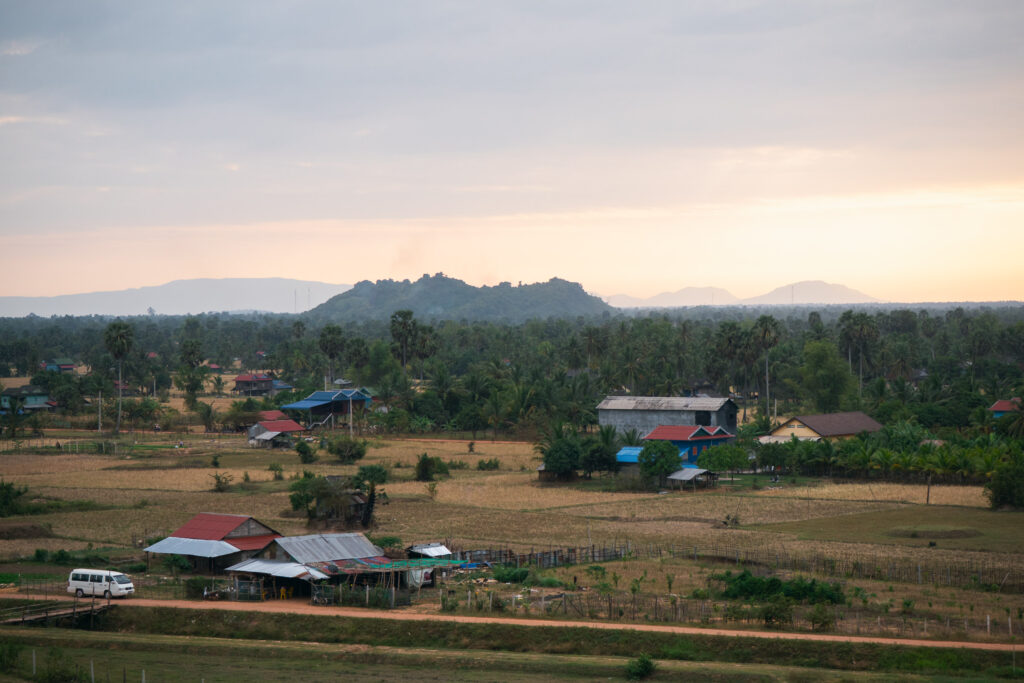 Rural landscape at sunset near Kampot, Cambodia, with scattered homes, palm trees, and forested hills in the distance.