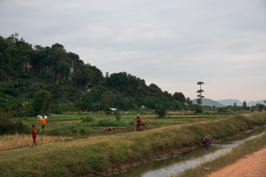 A rural landscape in Kampot with green fields, a tree-covered mountain, a canal, and scattered people working