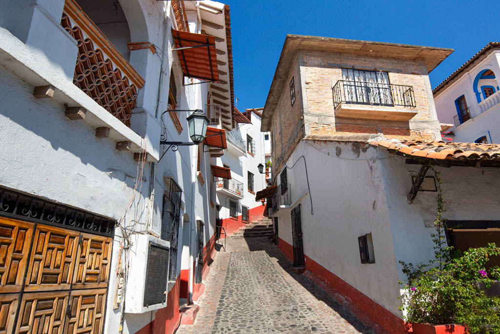 A narrow, cobblestone street in Taxco, Mexico, slopes uphill between whitewashed buildings with terracotta-tiled roofs