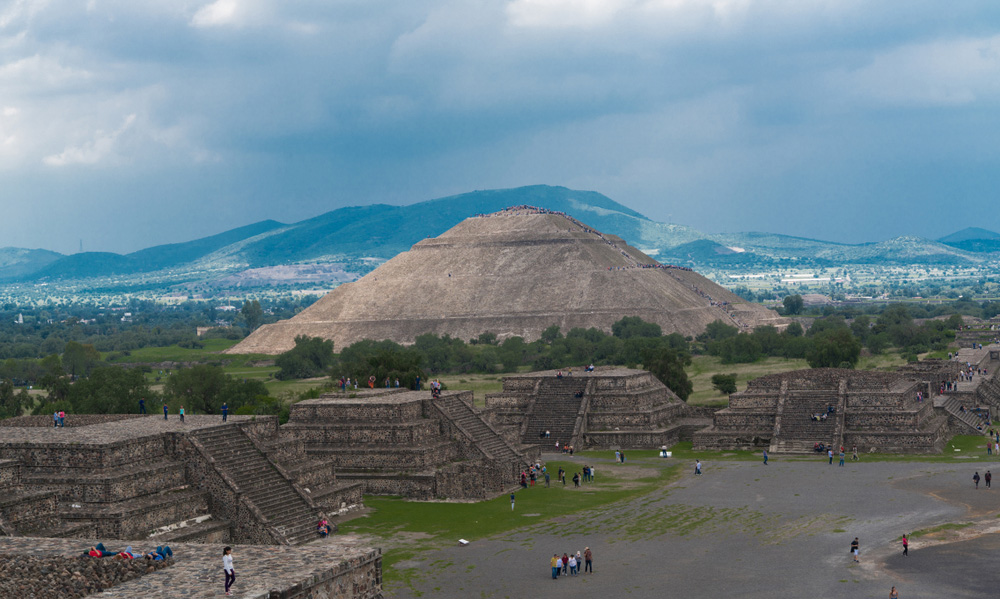 A wide shot of the Pyramid of the Sun in Teotihuacan, Mexico surrounded by trees and a mountain in the background