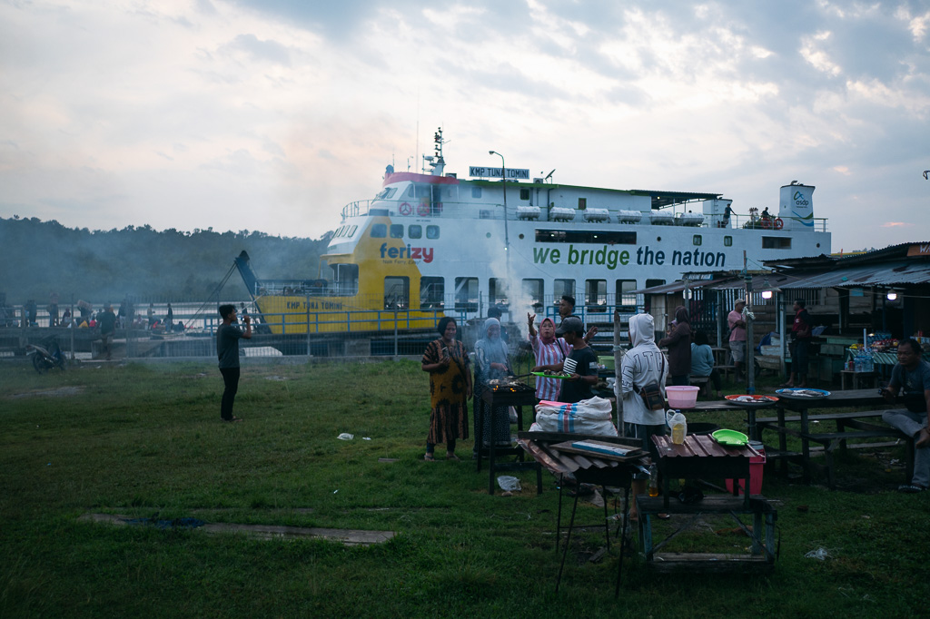 People gather around BBQ grills on a grassy area near a docked Ferizy ferry in the Togeans