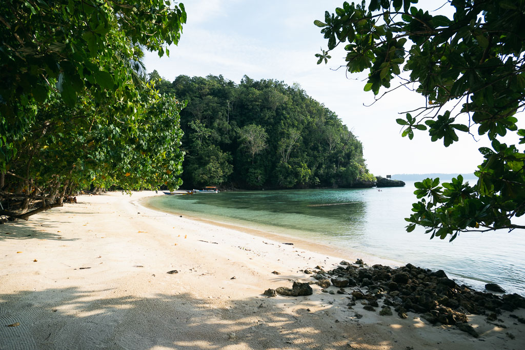 Secluded cove in the Togean Islands with white sand beach, turquoise water, lush greenery, and a small boat
