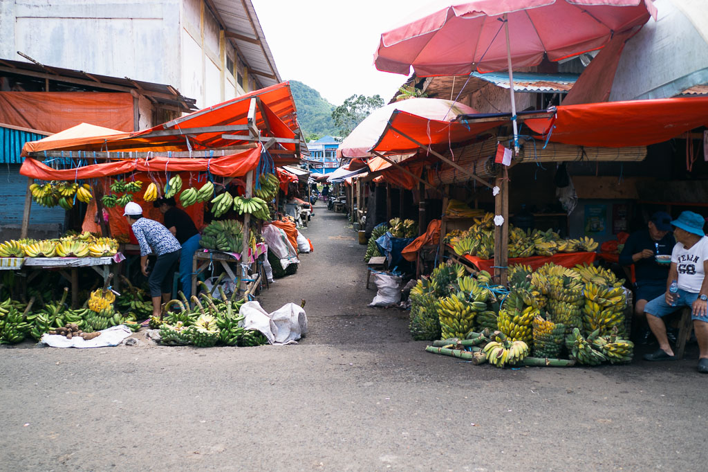 Busy outdoor market in Tomohon with stalls shaded by red and orange tarps, selling bananas and other produce
